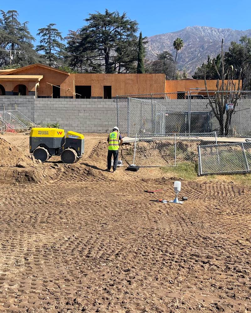 Titan technician in a safety vest on a graded residential building pad beside a trench roller, with a sand-cone density test set up in the foreground